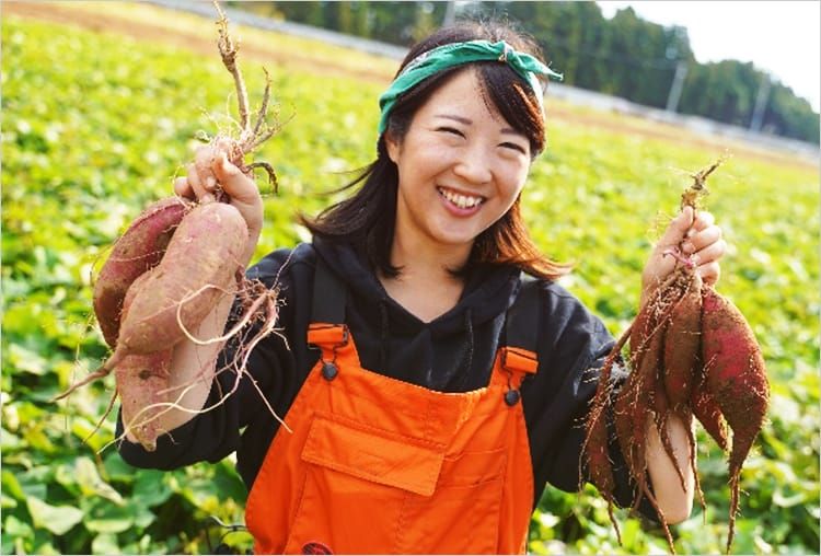 広大な畑を背景に収穫したばかりのサツマイモを両手に満面の笑みを浮かべている瀧澤芽衣さんの写真
